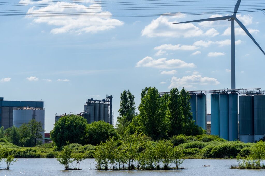 Scenic industrial landscape featuring a wind turbine, factories, and lush greenery by a body of water.